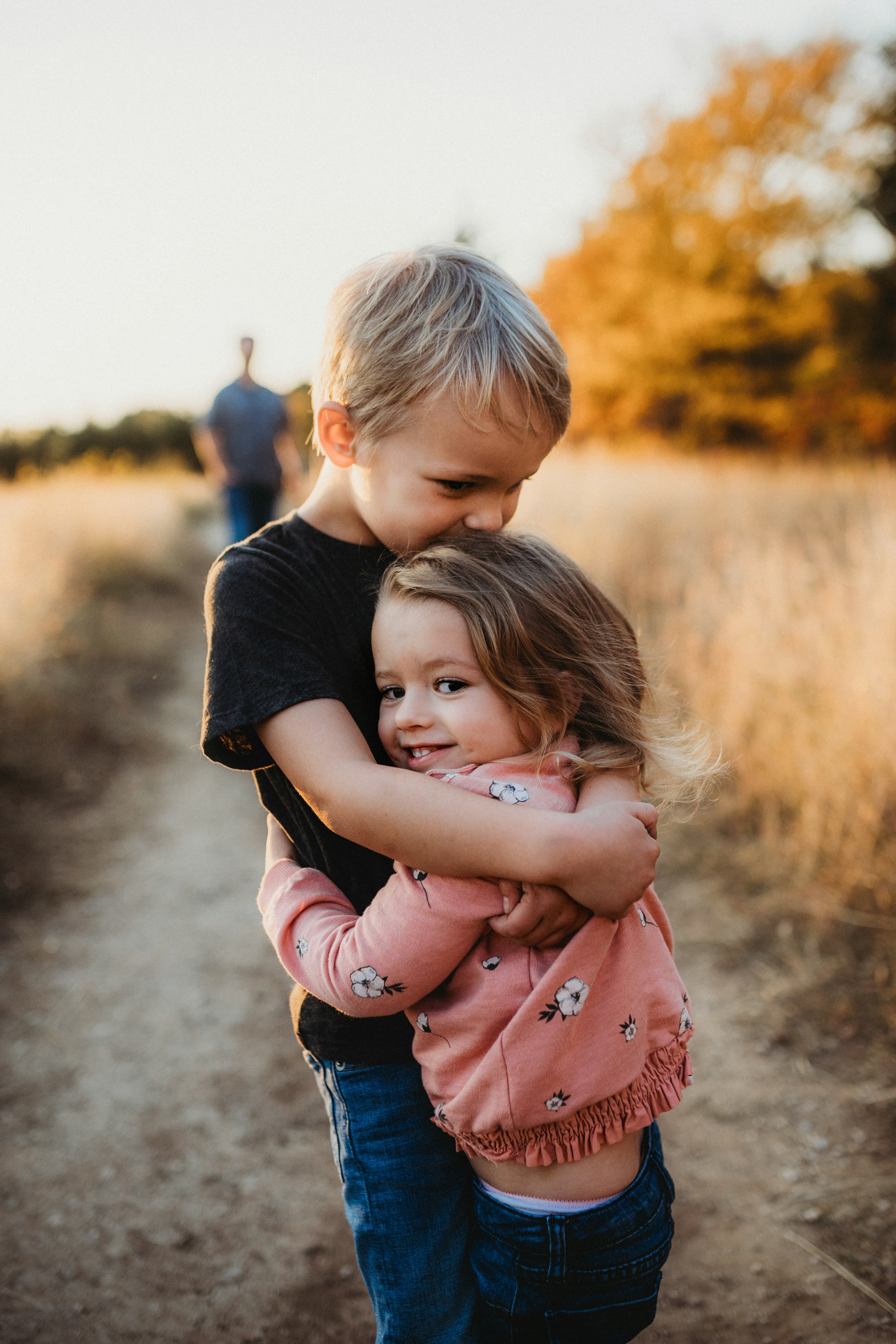A boy hugging his younger sister on a path with a parent in the background